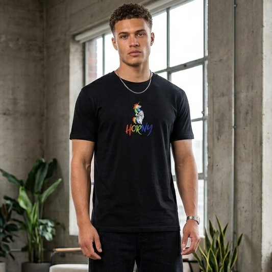 A man stands in a loft-style room with concrete walls and large windows, wearing a black graphic t-shirt with a colorful cartoon character and the word 'HORNY' printed on the chest, paired with black pants and white sneakers.