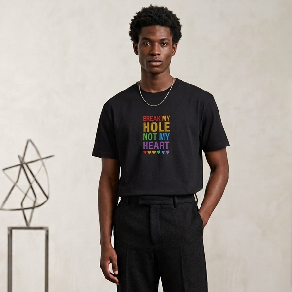 A man stands in a minimalist studio setting wearing a black graphic t-shirt with the text 'BREAK MY HOLE NOT MY HEART' in rainbow colors, paired with black trousers and black shoes.