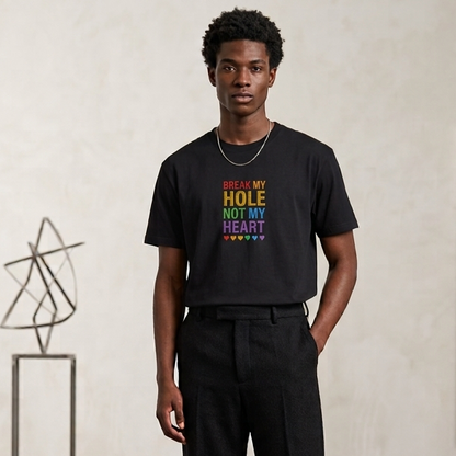 A man stands in a minimalist studio setting wearing a black graphic t-shirt with the text 'BREAK MY HOLE NOT MY HEART' in rainbow colors, paired with black trousers and black shoes.