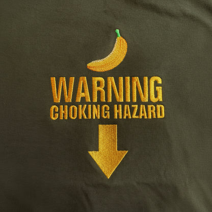 Close-up of embroidered text reading 'WARNING CHOKING HAZARD' in gold thread, with a small banana graphic above and a large downward arrow below, on a dark fabric background.