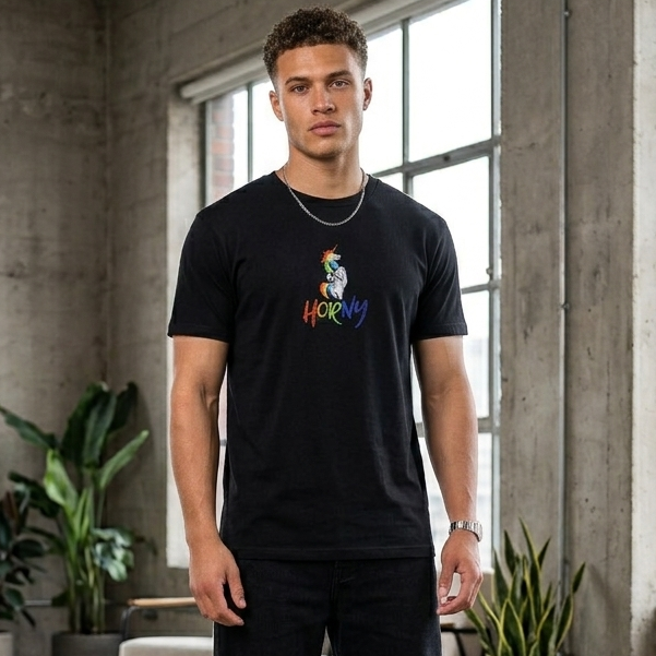 A man stands in a loft-style room with concrete walls and large windows, wearing a black graphic t-shirt with a colorful cartoon character and the word 'HORNY' printed on the chest, paired with black pants and white sneakers.