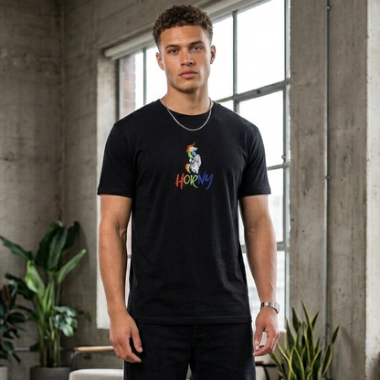 A man stands in a loft-style room with concrete walls and large windows, wearing a black graphic t-shirt with a colorful cartoon character and the word 'HORNY' printed on the chest, paired with black pants and white sneakers.